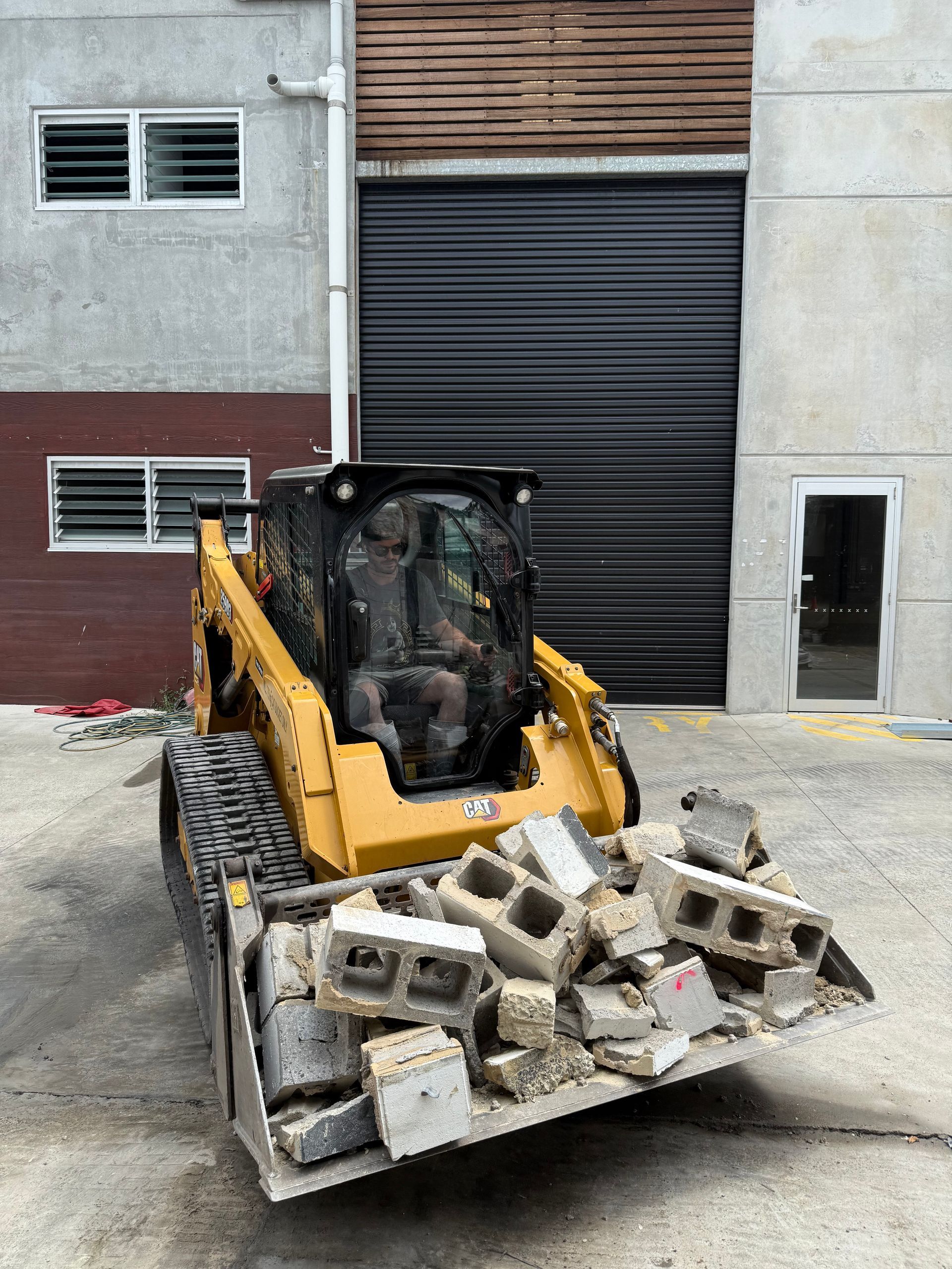Yellow skid steer loader carrying concrete blocks in a work area near a warehouse.