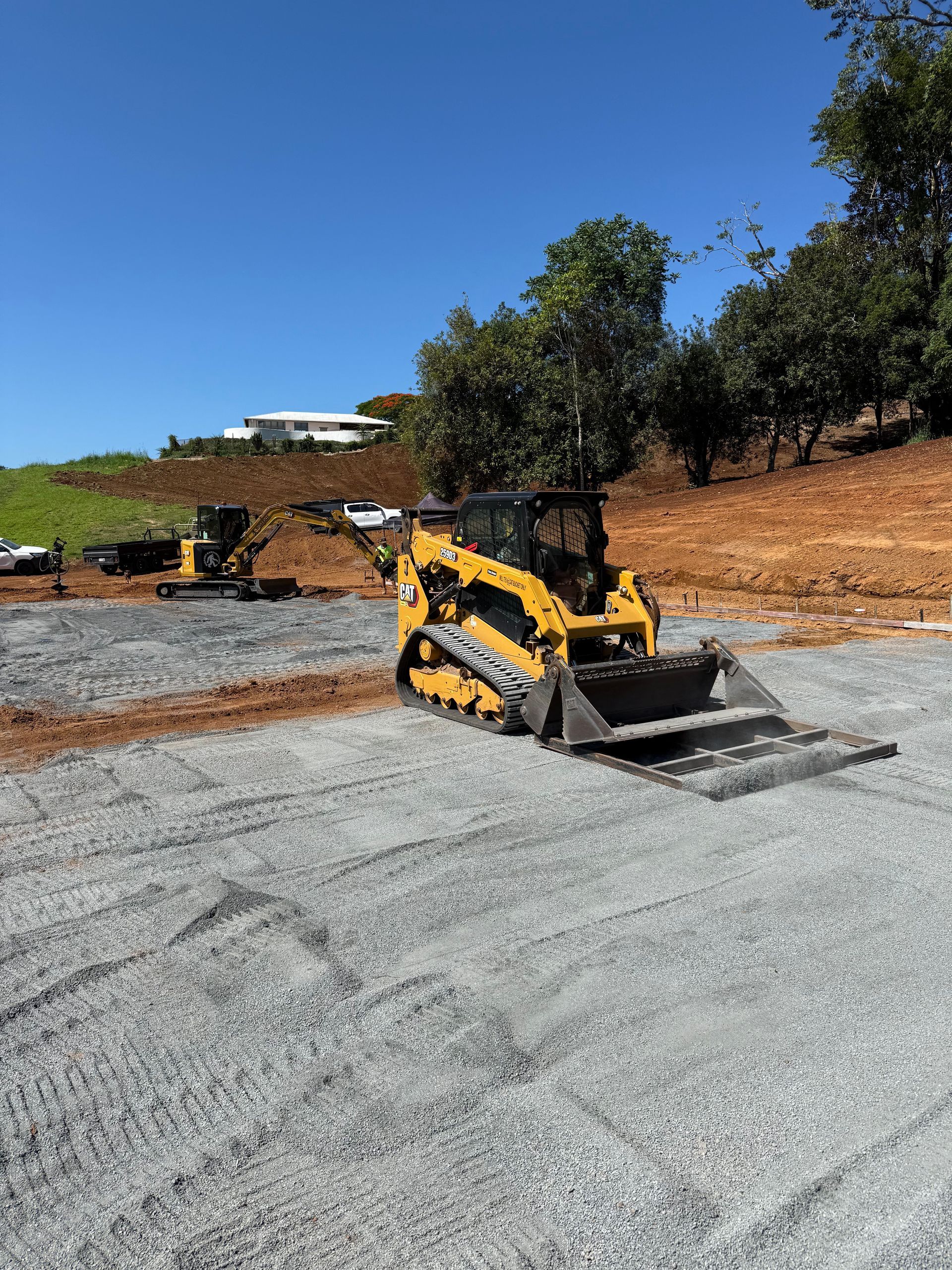 Yellow skid steer with bucket on a gravel surface, another in the background. Construction site, bright blue sky.
