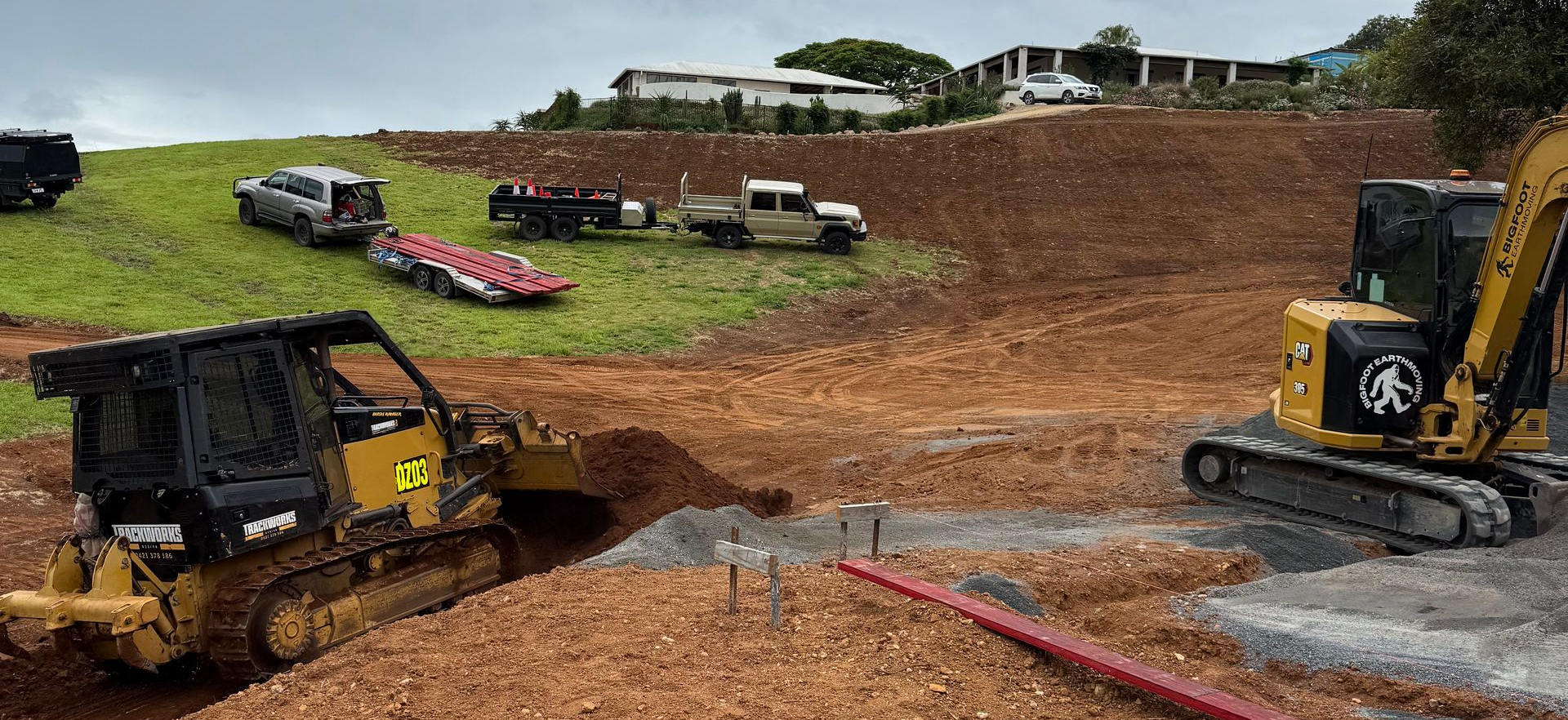 Construction site with a bulldozer and excavator grading a hillside. Cars and trucks are parked nearby.