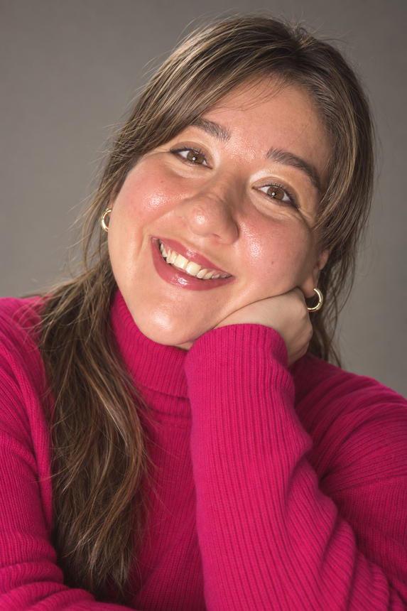 Smiling person in a bright pink sweater, resting their chin on their hand against a gray background.