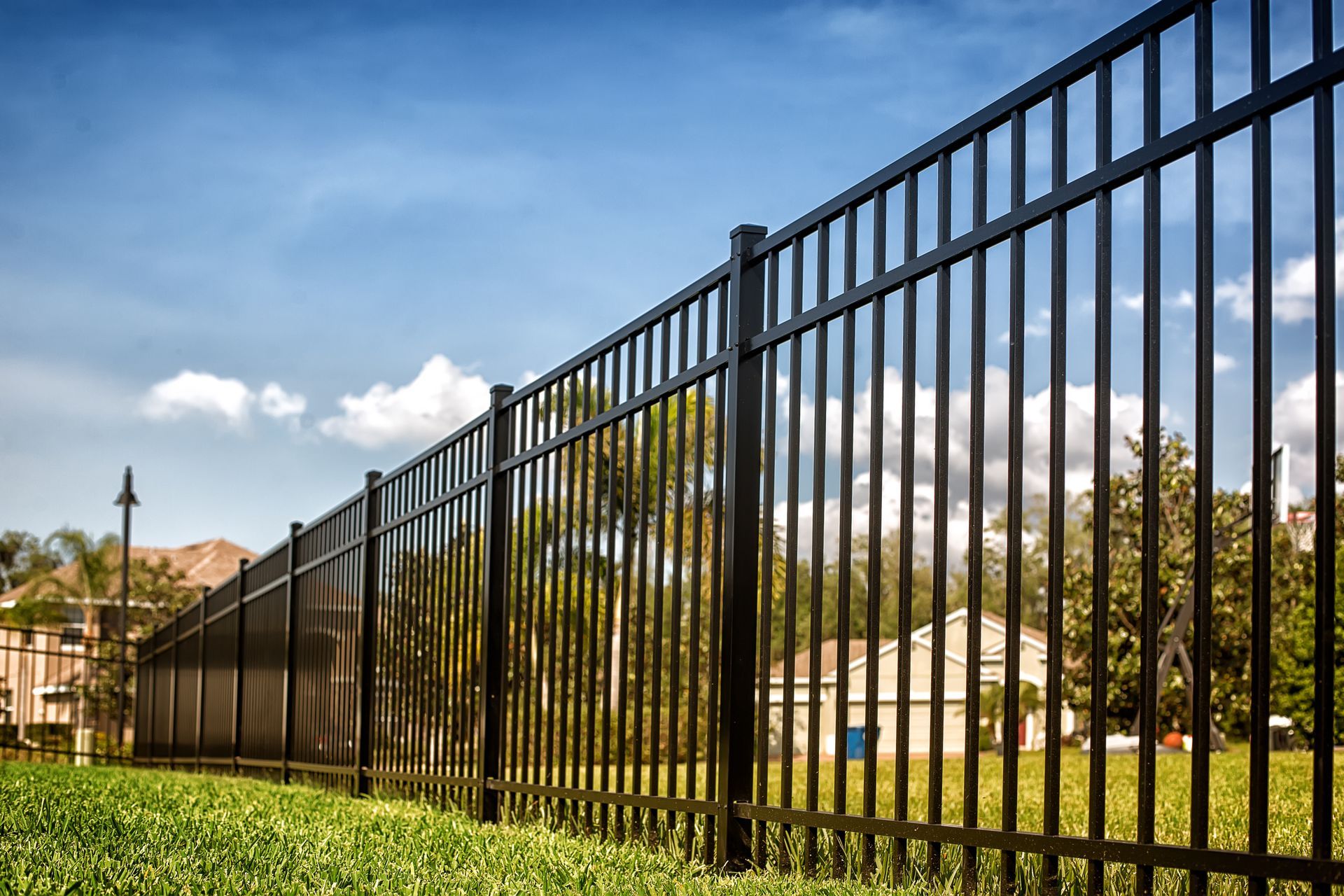 Black metal fence beside a grassy field under a blue sky with clouds and trees