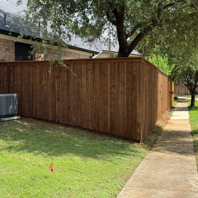 Wooden privacy fence along a grassy yard beside a concrete sidewalk under a large tree