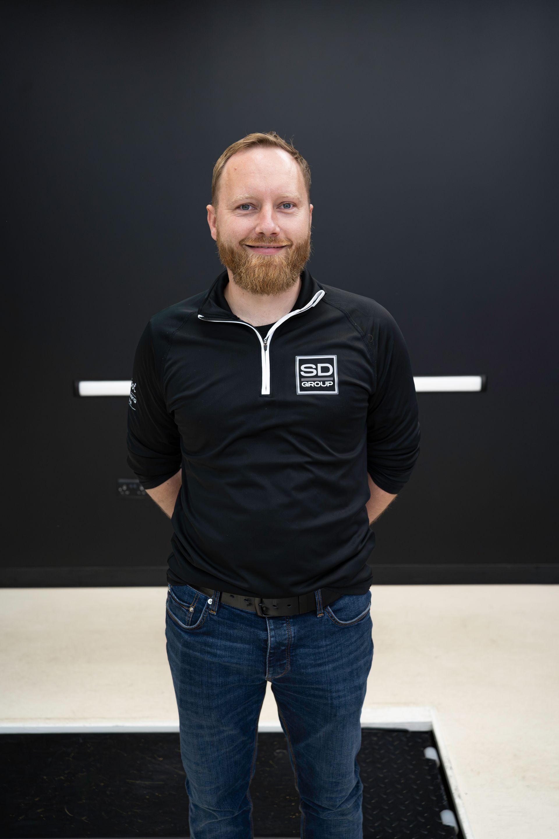 Man with a beard in a black shirt and jeans stands in front of a black wall.