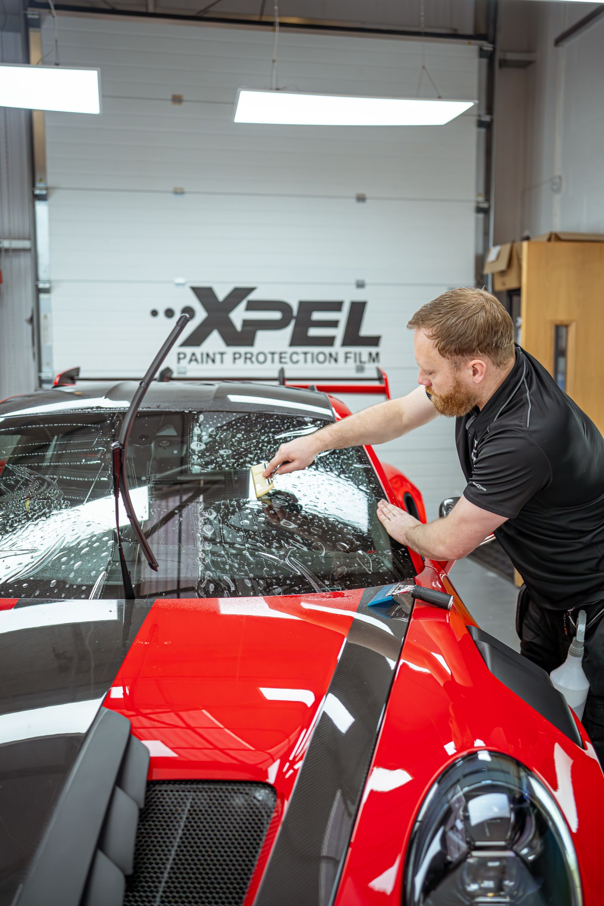 A man is cleaning the windshield of a red car in a garage.