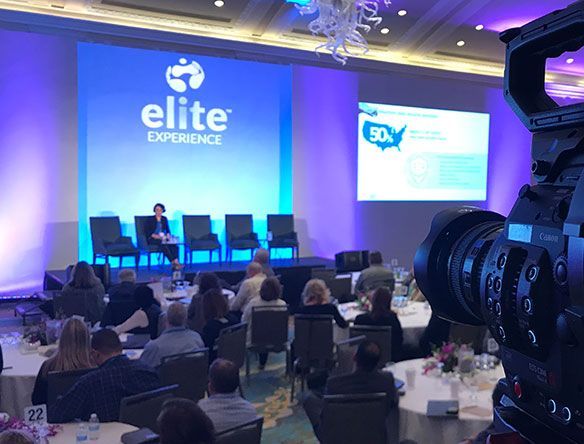 Conference panel on a blue-lit stage with seated audience and a camera filming from the foreground