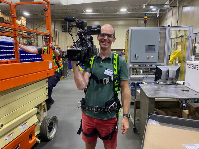 Smiling person holding a camera in an industrial workshop beside large machinery