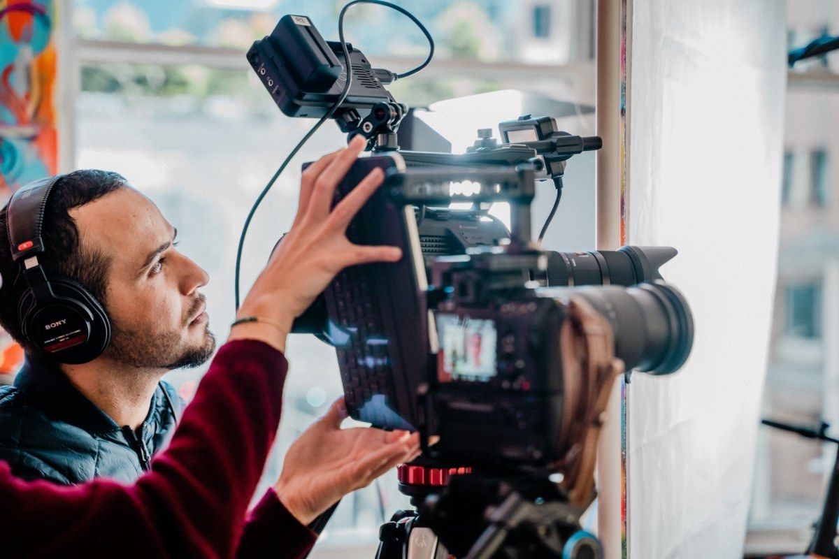 Camera operator adjusting a professional video camera on a tripod indoors near a bright window