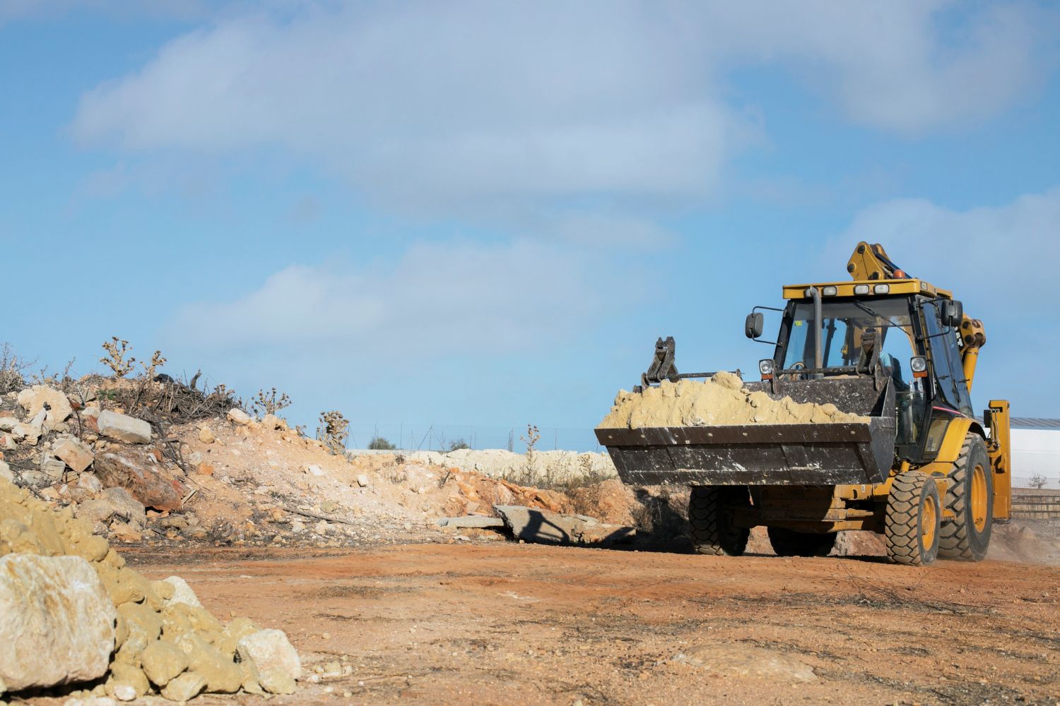 A yellow backhoe loader carries a bucket full of dirt across a rocky, construction site under a blue sky.