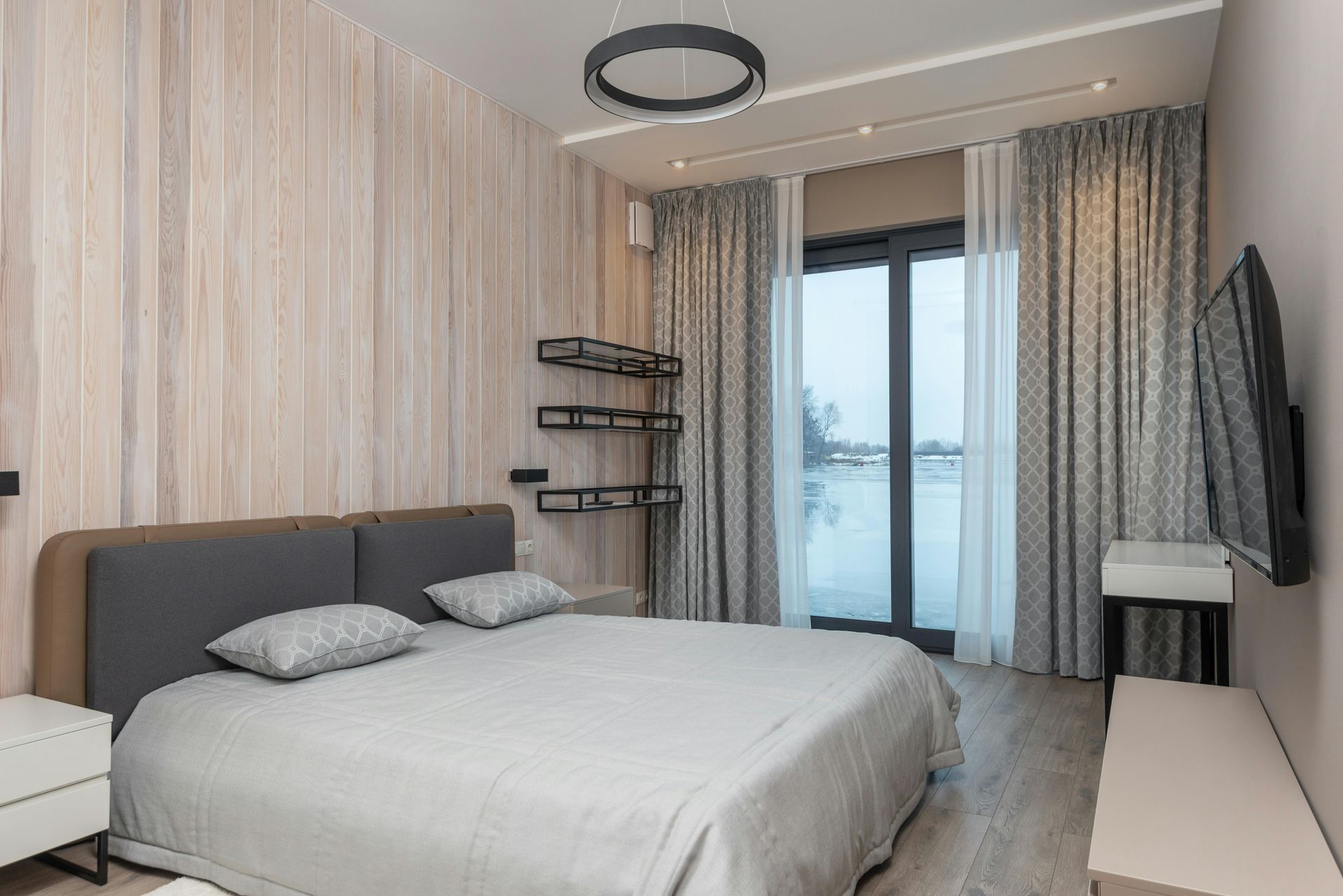 A modern bedroom with a wood-paneled accent wall, a gray bed, wall-mounted shelves, and floor-to-ceiling curtained windows.