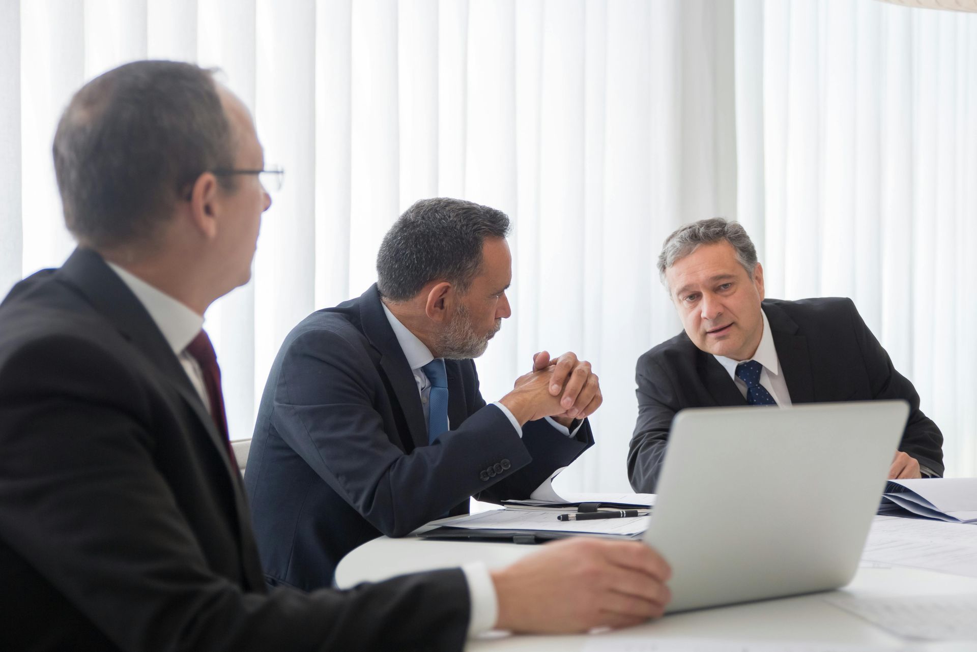 Three professionals in suits sit at a table, discussing business while looking at a laptop in a bright, modern office.