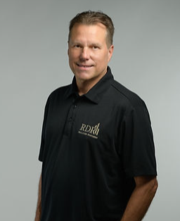 Man in a black polo shirt with a logo, standing against a gray studio background.