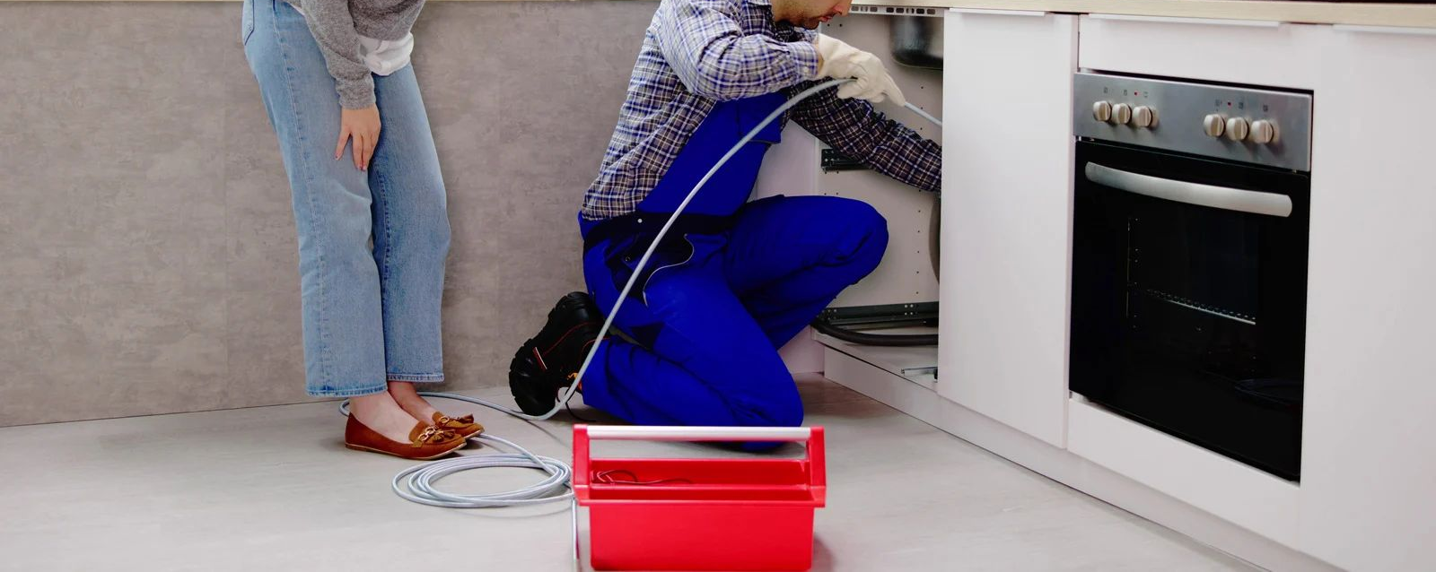 Person kneeling by a stove, painting kitchen cabinets while another stands nearby with tools and paint tray