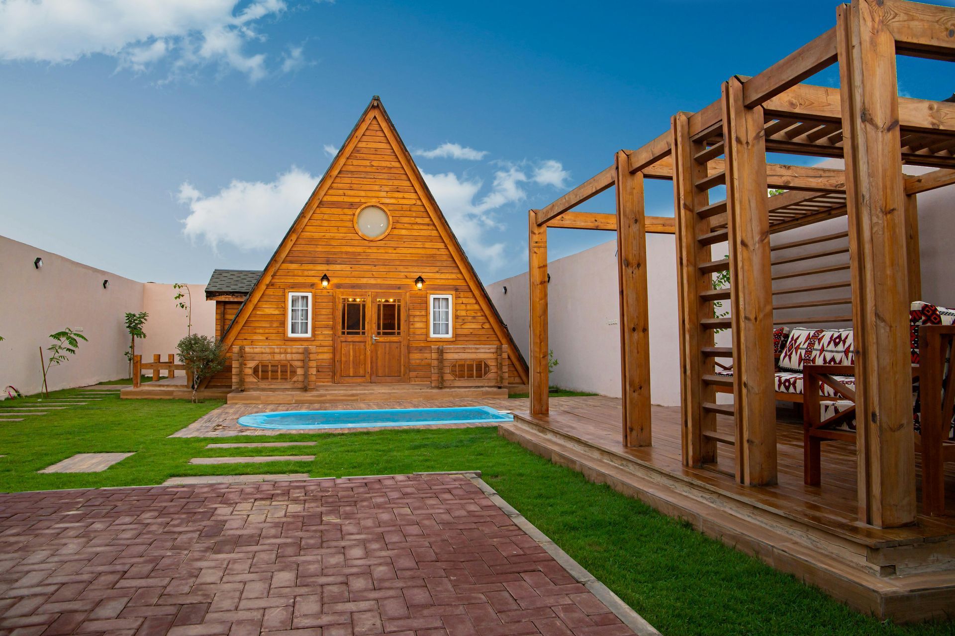 Wooden A-frame cabin with small pool, patio, and pergola, set in a yard with green grass against a clear blue sky.