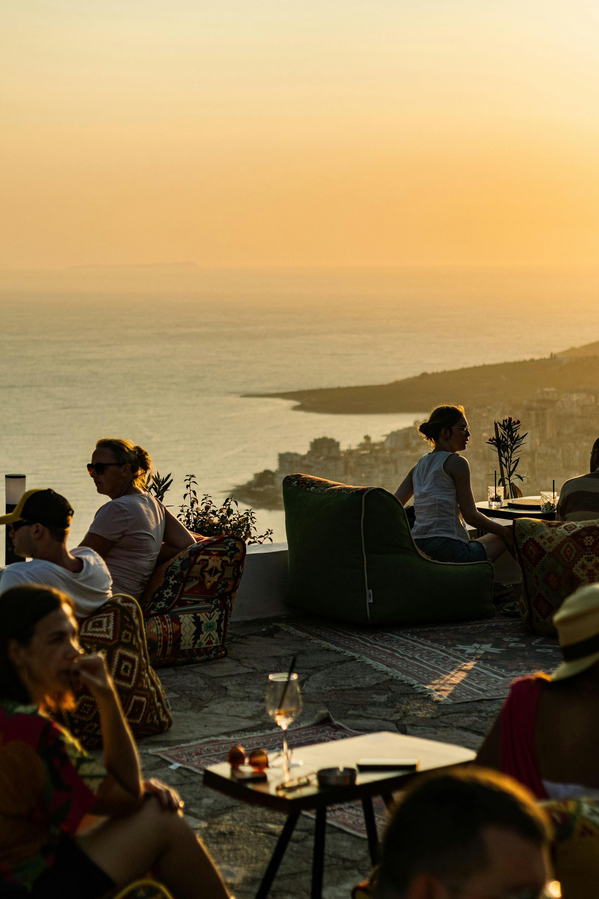 People sit on a terrace overlooking the sea at sunset, enjoying drinks.