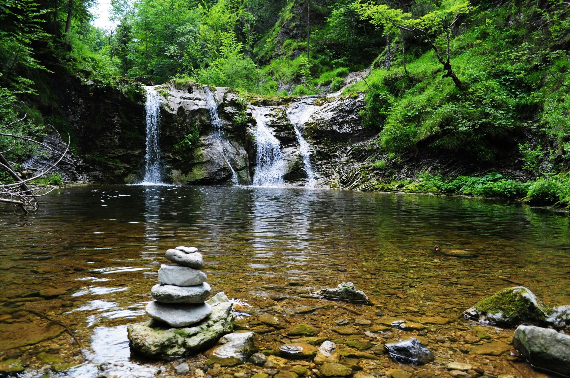 Waterfall cascading into a clear pool, with stacked stones in the foreground and lush green foliage.