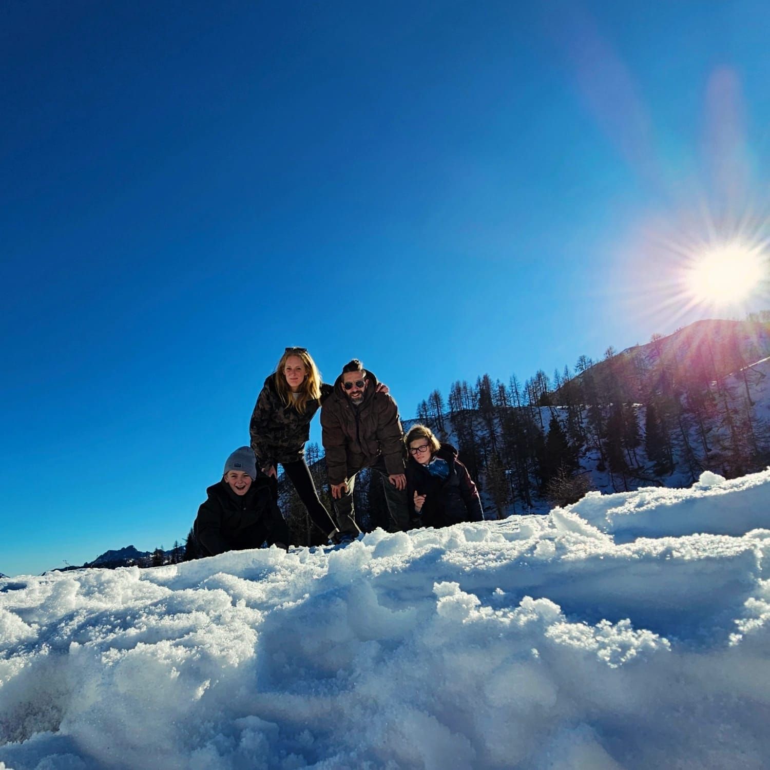 Vier mensen poseren in de sneeuw, met een felle zon aan hun zijde. Blauwe lucht, besneeuwde grond en beboste heuvels op de achtergrond.