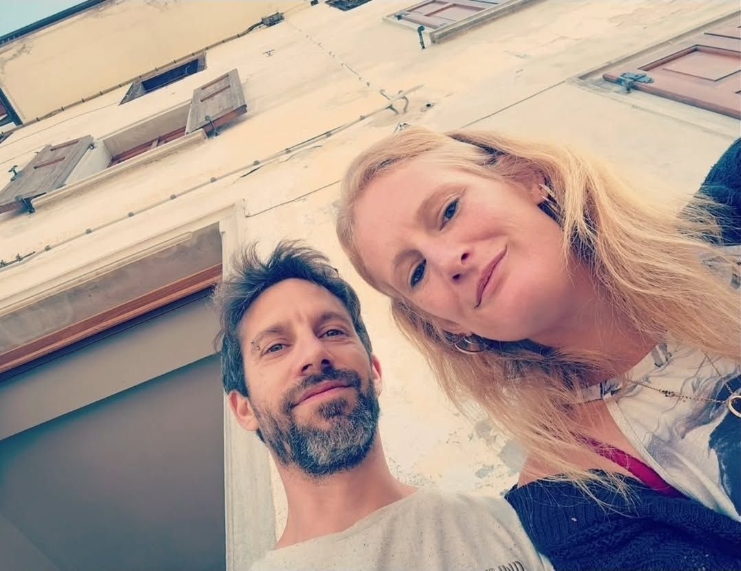 Een man met een baard en een vrouw met blond haar maken een selfie, met een wit gebouw op de achtergrond.