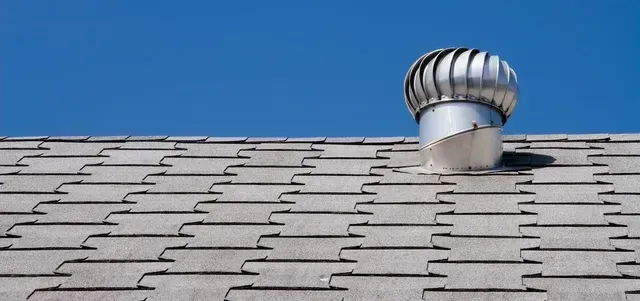 A roof with gray shingles and a spinning vent against a clear blue sky.