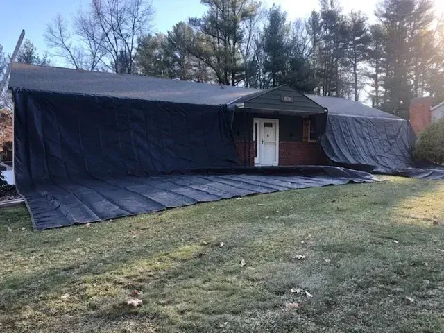 House covered with a large black tarp, likely for roof repair. Tarp extends onto the lawn.