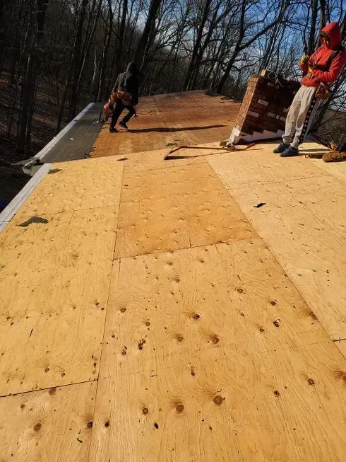 Two workers on a plywood roof installing material under a clear sky.