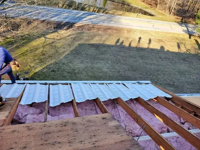 Man installing metal roofing panels, roof partially exposed with pink insulation. Sunny outdoor setting.