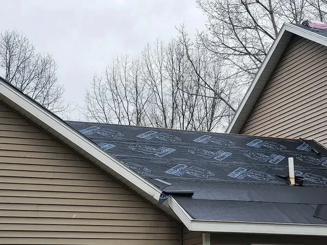 Roof with black underlayment partially covering the shingles on a house with tan siding.