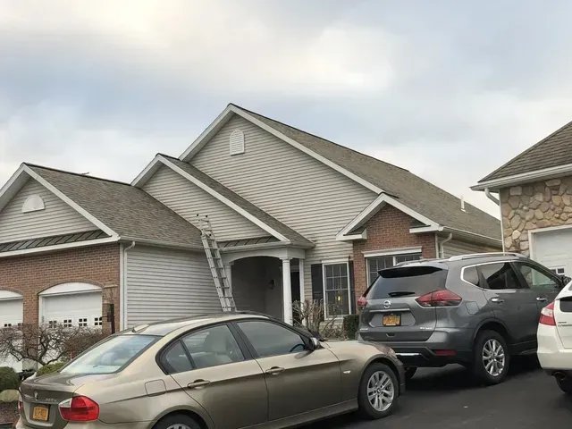 A house with a car parked in the driveway and a ladder leaning against the roof. Overcast day.