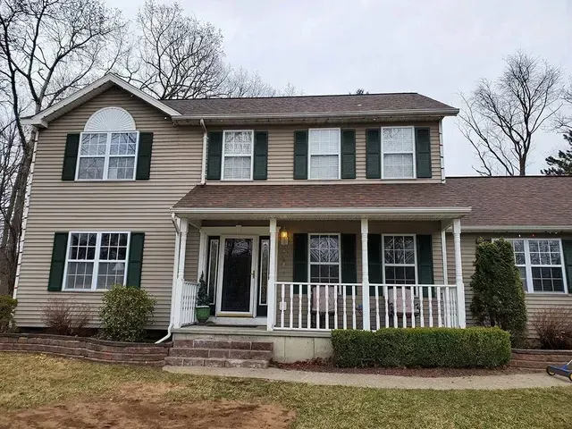 Tan two-story house with green shutters, white trim, and a porch. Overcast sky, trees in the background.
