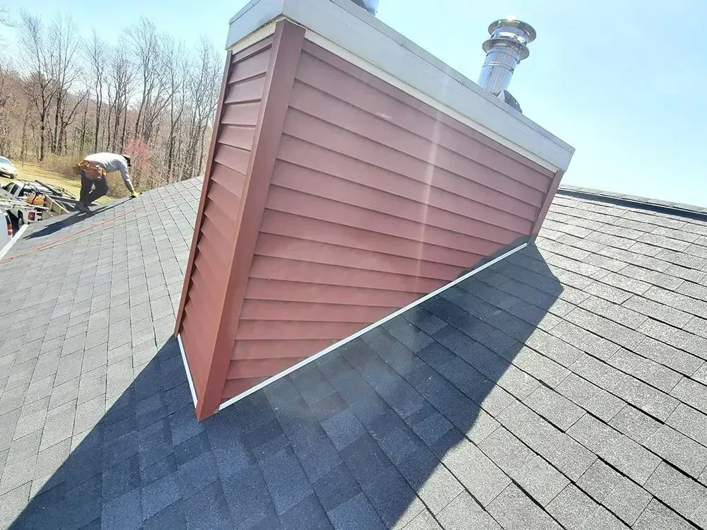 Chimney with red siding on a gray shingle roof under a clear, sunny sky; person working on the roof.