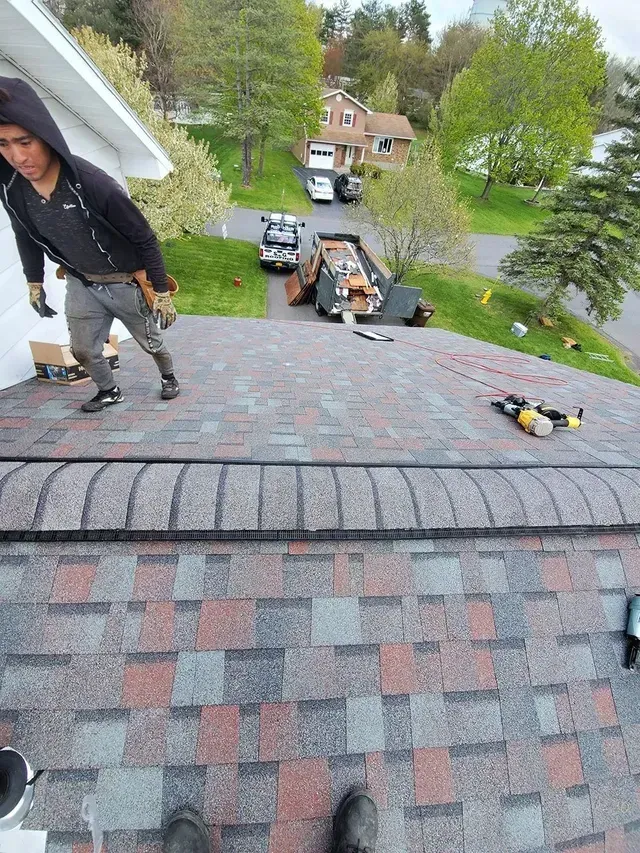 Roofer on a shingled roof, next to tools. Man in a hoodie. Cars and trees in the background.