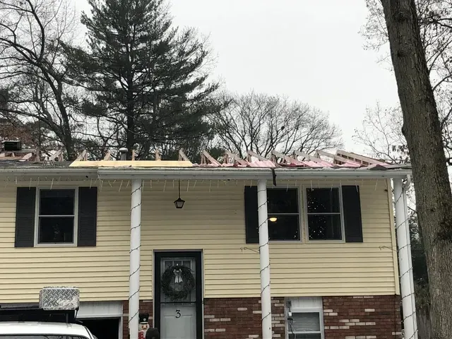 House with roof partially removed during renovation. Beige siding, black shutters, brick base.