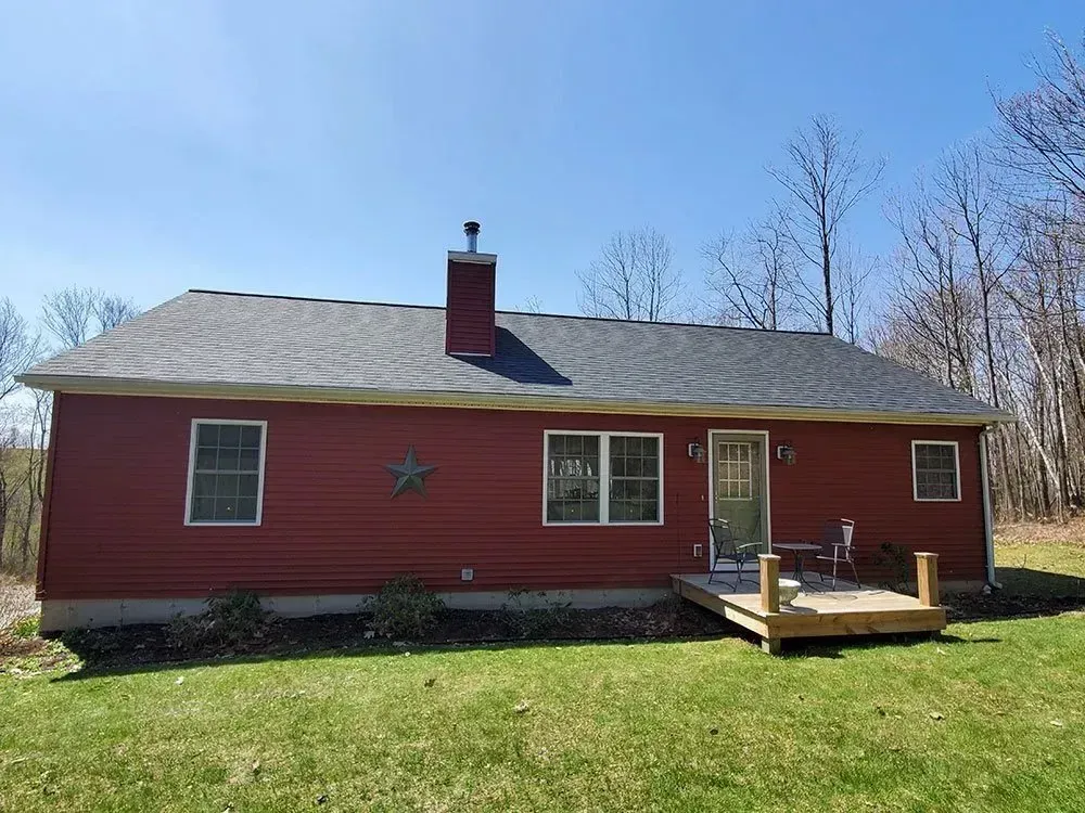 Red-sided ranch house with a wooden deck and chimney, set against a backdrop of trees and a blue sky.