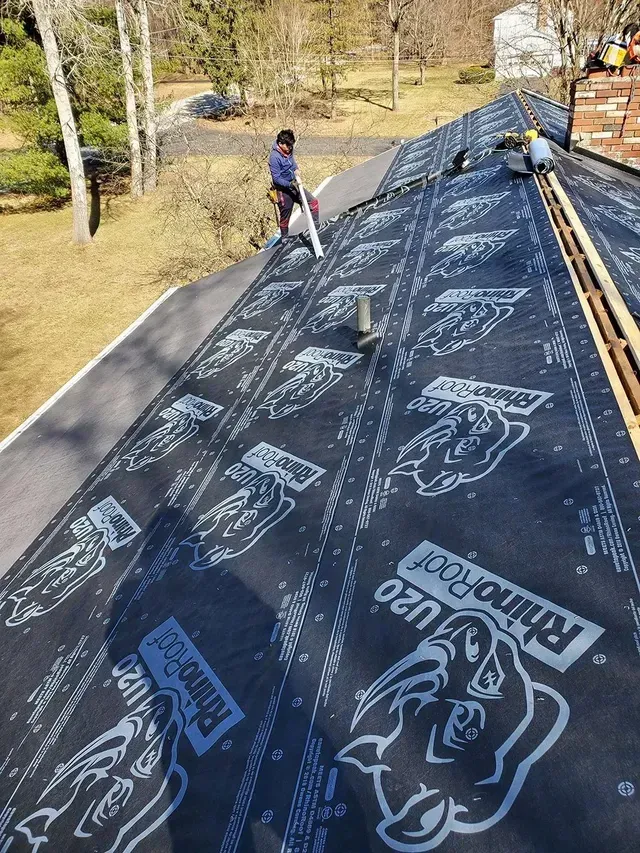 Roofers installing RhinoRoof underlayment on a house roof. Blue and black material, sunny day.