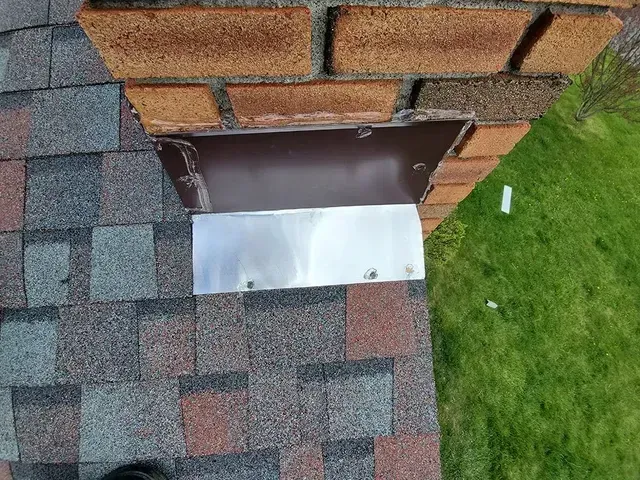 Chimney flashing with metal cover, mounted on brick and asphalt shingles, over green grass.