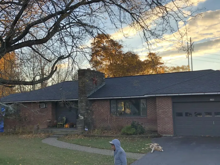 Brick ranch house with black roof, chimney, and garage. Man walking on driveway. Fall foliage and cloudy sky.