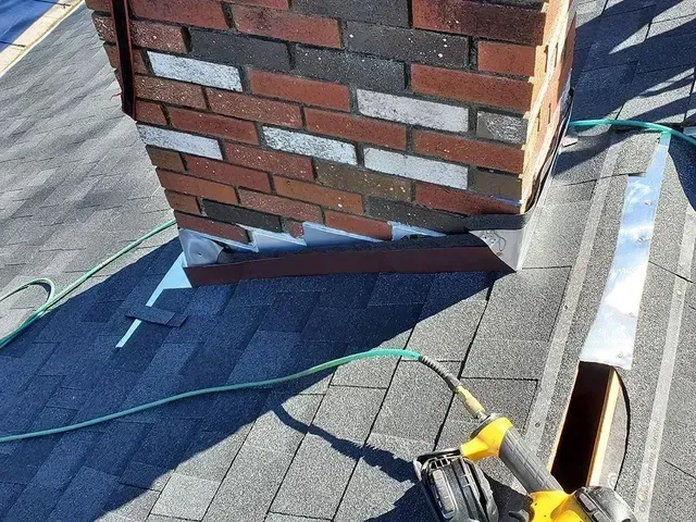 Chimney with metal flashing and roofing work in progress; brick and gray shingles.