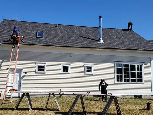 Roofers working on a house with a dark gray shingled roof, a chimney, and windows under a blue sky.