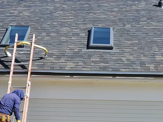 Person on a ladder, working on a roof with two skylights. The roof is dark with gray/brown shingles.