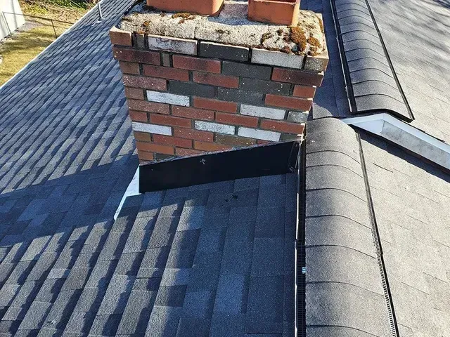 Chimney on a dark-shingled roof with flashing, viewed from above on a sunny day.