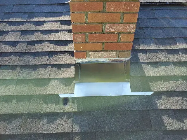 Brick chimney on a shingled roof with metal flashing around the base.