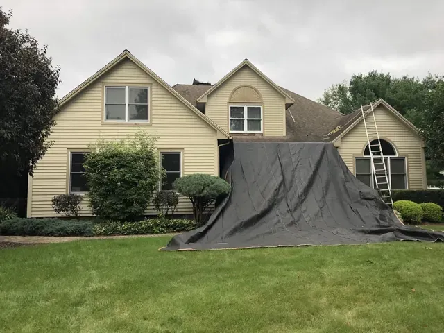House with tarp covering roof, ladder leans against it. Cloudy day.