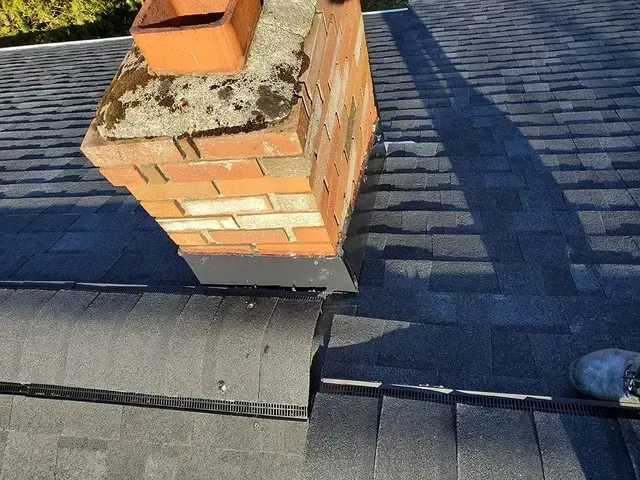 Brick chimney on a dark shingle roof with flashing at the base, angled view, outdoor setting.