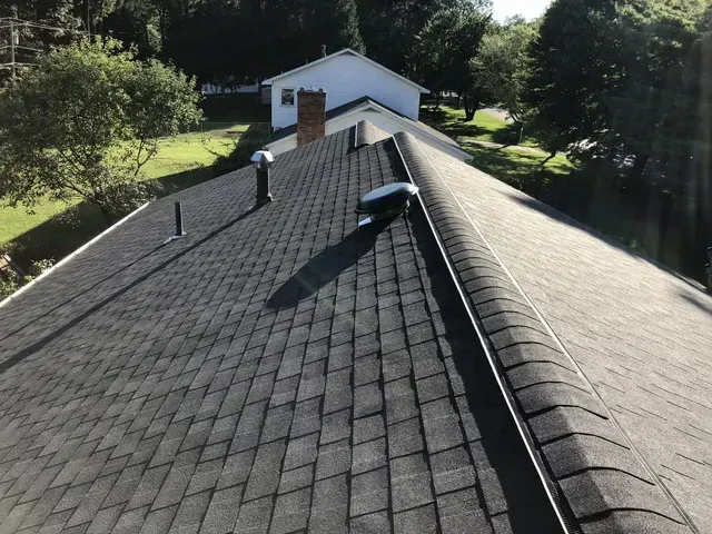 A dark gray asphalt shingle roof on a house with a surrounding green landscape and blue sky.