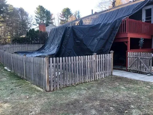Black tarp covers a house roof and deck, secured by a wooden fence in a grassy yard.