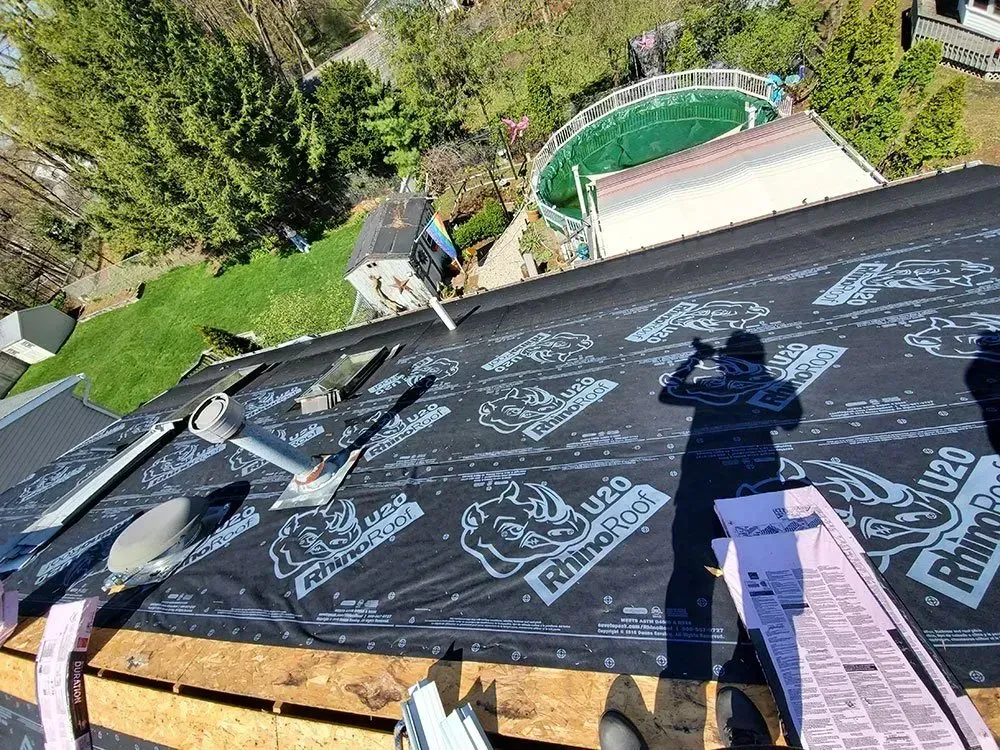 Roofer working on a house roof, covered in black underlayment, tools nearby, sunny day.