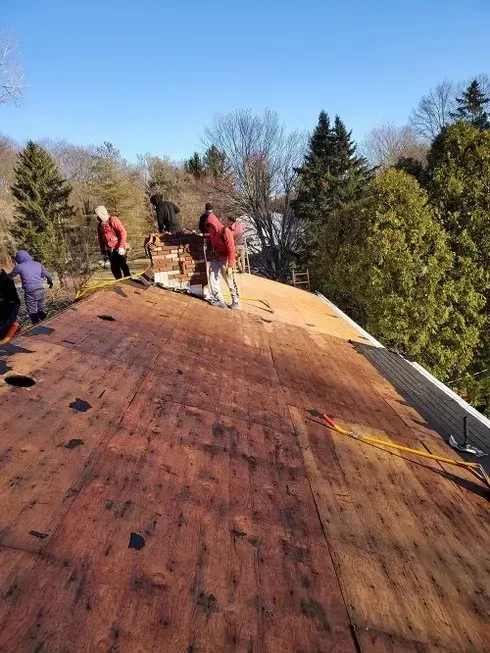 People working on a roof, removing shingles. Blue sky, trees in background.
