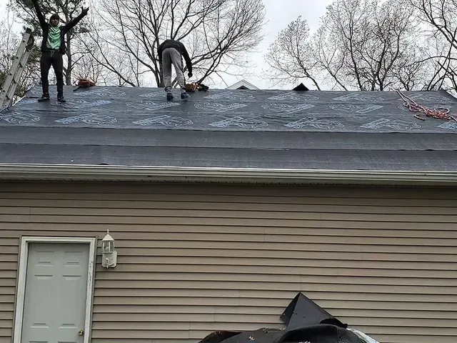 Two people on a dark roof, one with arms raised. Overcast day. House with white door and beige siding below.