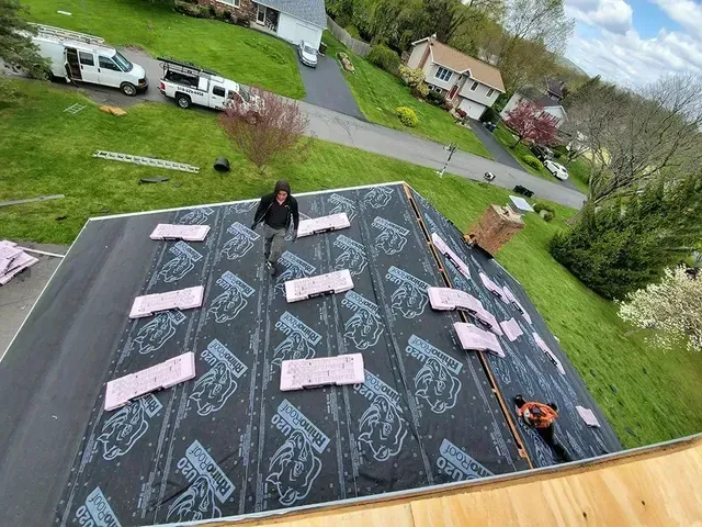 Roofer working on a dark roof with pink insulation pieces. View includes houses, a van, and green grass.