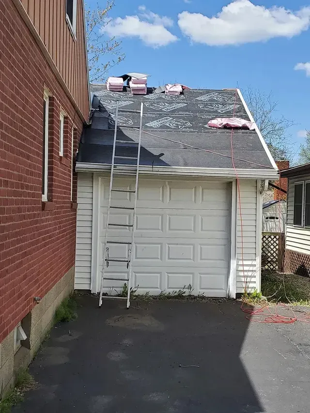 A ladder leans against a garage with a partially torn-off roof; pink insulation and asphalt shingles are visible.