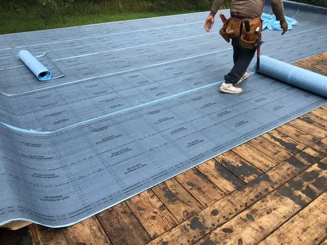 Person rolling out blue roofing underlayment on weathered wooden boards.